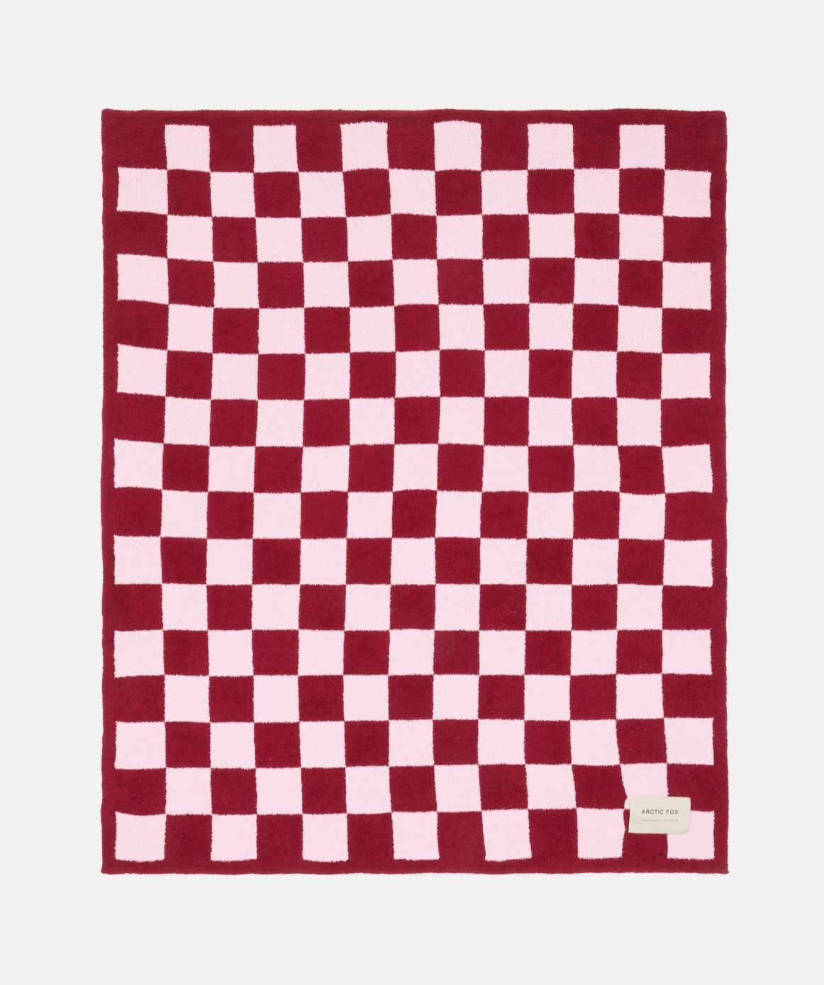 A rectangular knit blanket with a red and light pink checkerboard pattern, laid flat against a light gray background. A small white brand label is visible in the bottom right corner.