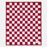 A rectangular knit blanket with a red and light pink checkerboard pattern, laid flat against a light gray background. A small white brand label is visible in the bottom right corner.