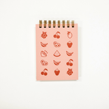 A pink spiral-bound notepad featuring red fruit illustrations, photographed against a clean white background.