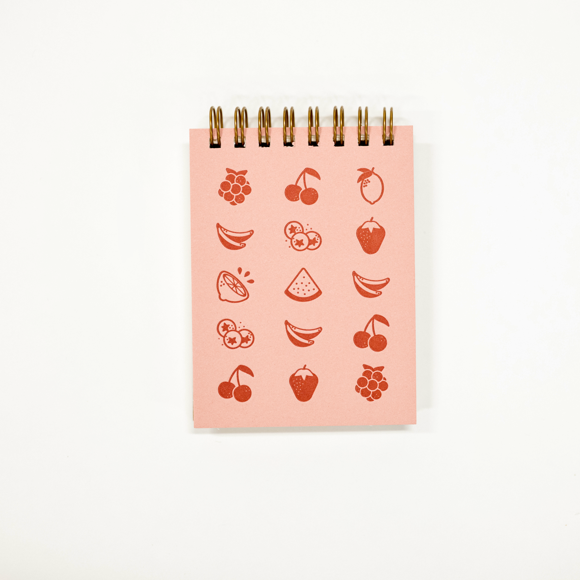 A pink spiral-bound notepad featuring red fruit illustrations, photographed against a clean white background.