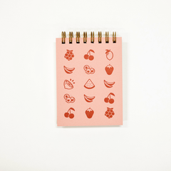 A pink spiral-bound notepad featuring red fruit illustrations, photographed against a clean white background.