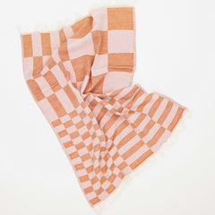 A woven kitchen towel with a light pink and deep pink checkerboard pattern and fringed edges, laid out on a clean white background.