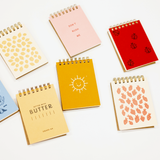 Seven spiral-bound notepads in assorted pastel and bright colors are arranged on a clean white background. Each notebook has a unique cover design, including patterns of leaves, strawberries, hearts, and illustrations, with one yellow notebook featuring a smiling sun and another in yellow-beige reading 'BUTTER'.