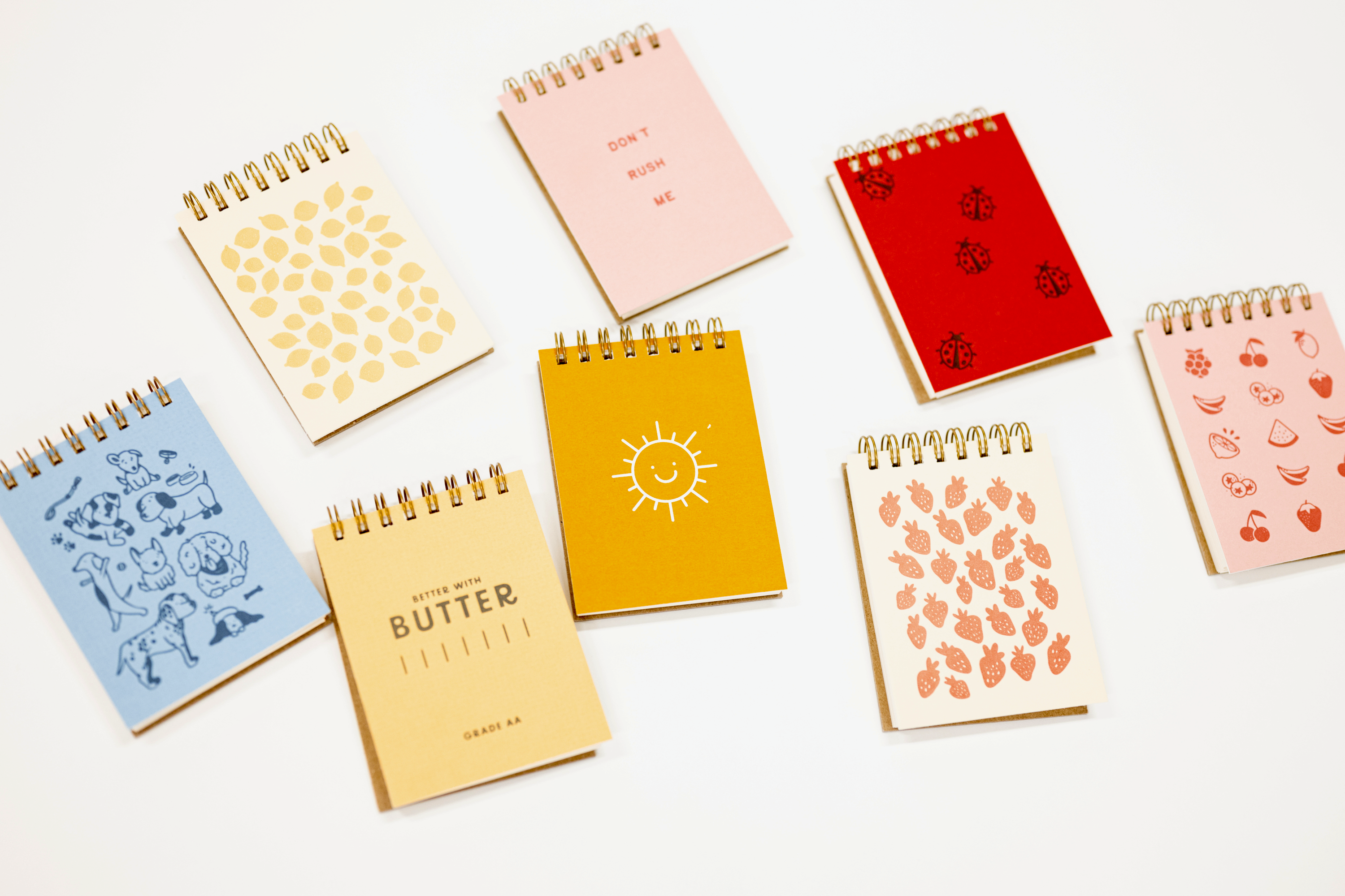 Seven spiral-bound notepads in assorted pastel and bright colors are arranged on a clean white background. Each notebook has a unique cover design, including patterns of leaves, strawberries, hearts, and illustrations, with one yellow notebook featuring a smiling sun and another in yellow-beige reading 'BUTTER'.