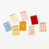 Seven small spiral-bound notepads in assorted pastel and bright colors, arranged on a clean white background. The covers feature different patterns and text, including 'BUTTER FLIGHT', a sun illustration, strawberries, cherries, and hearts.