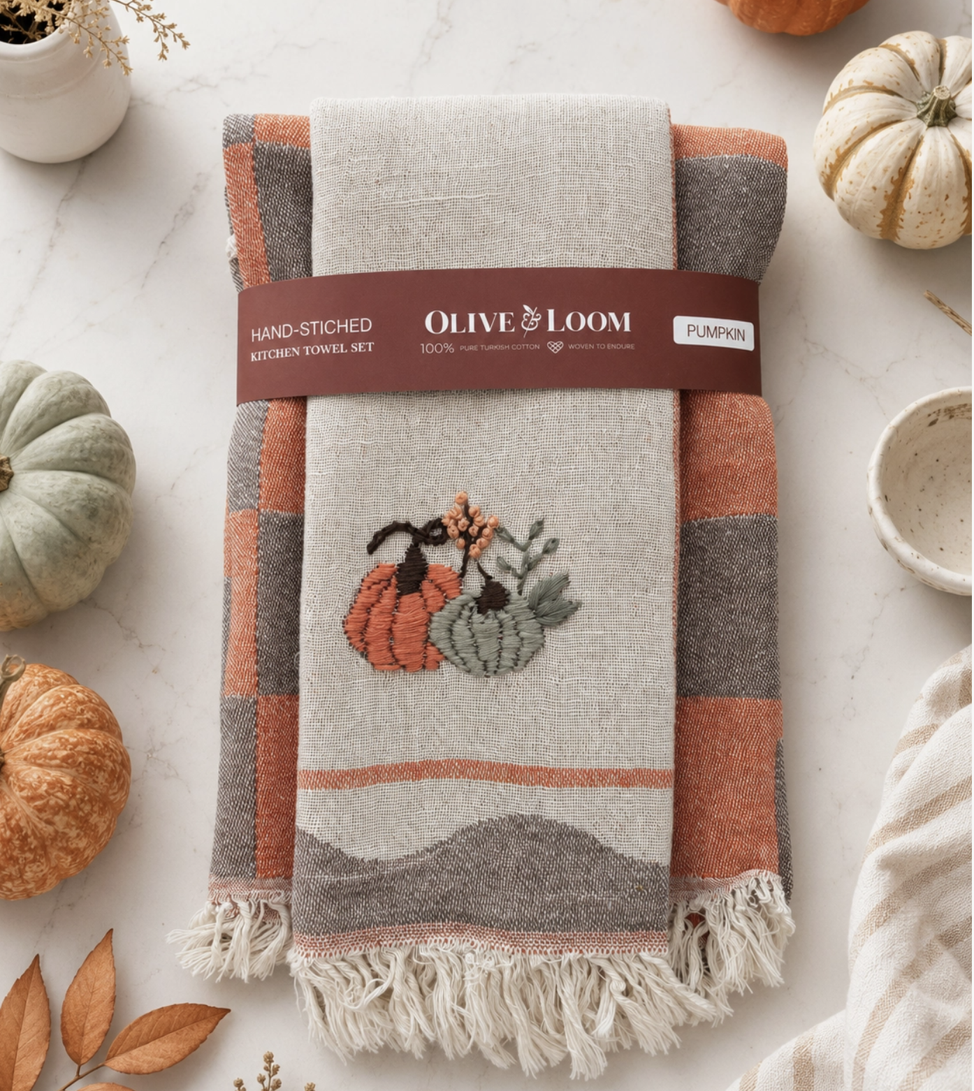 A folded kitchen towel set wrapped in a paper band labeled 'Olive Loom Pumpkin' and 'Hand-Stitched Kitchen Towel Set,' placed on a light marble surface with small pumpkins and autumn leaves around it. The towel has a cream base with orange, taupe, and gray plaid sections and a stitched pumpkin motif near the center.