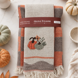 A folded kitchen towel set wrapped in a paper band labeled 'Olive Loom Pumpkin' and 'Hand-Stitched Kitchen Towel Set,' placed on a light marble surface with small pumpkins and autumn leaves around it. The towel has a cream base with orange, taupe, and gray plaid sections and a stitched pumpkin motif near the center.