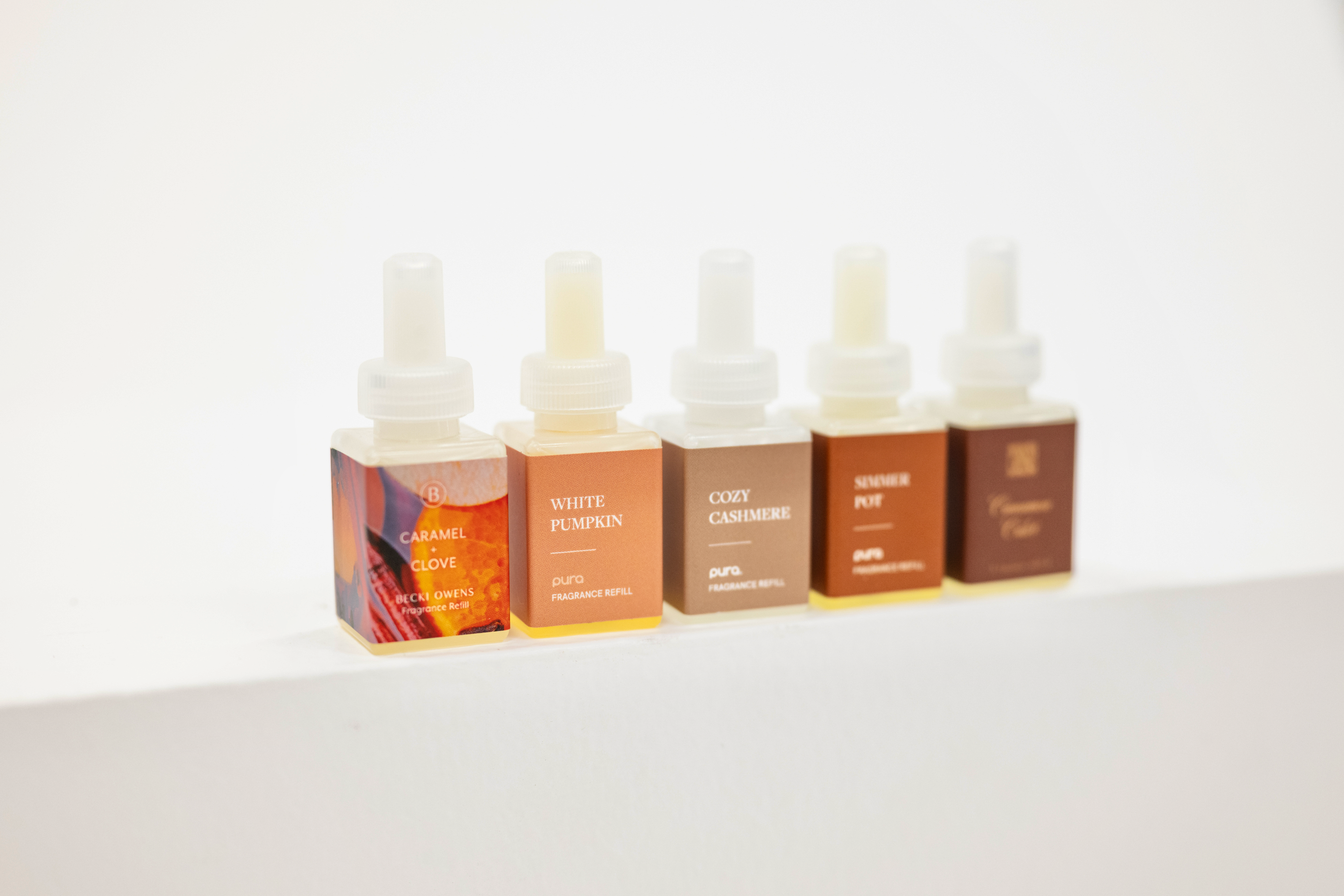 Fall fragrances for Pura. Five small rectangular fragrance oil bottles with white dropper tops lined up on a white surface against a white background. The bottles have warm-toned labels in orange, brown, and beige, with visible text including 'Caramel Clove', 'White Pumpkin'
