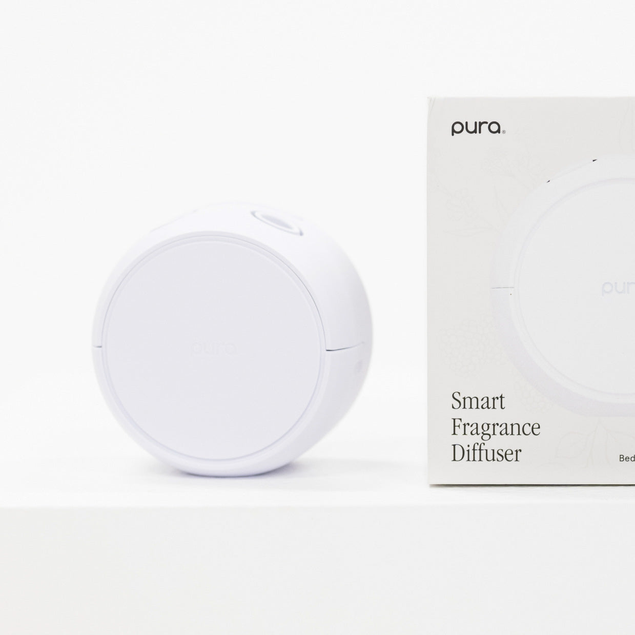 A white round smart fragrance diffuser sits beside its white retail box on a clean white background. The box shows the Pura brand name and the text 'Smart Fragrance Diffuser'.