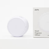 A white round smart fragrance diffuser sits beside its white retail box on a clean white background. The box shows the Pura brand name and the text 'Smart Fragrance Diffuser'.