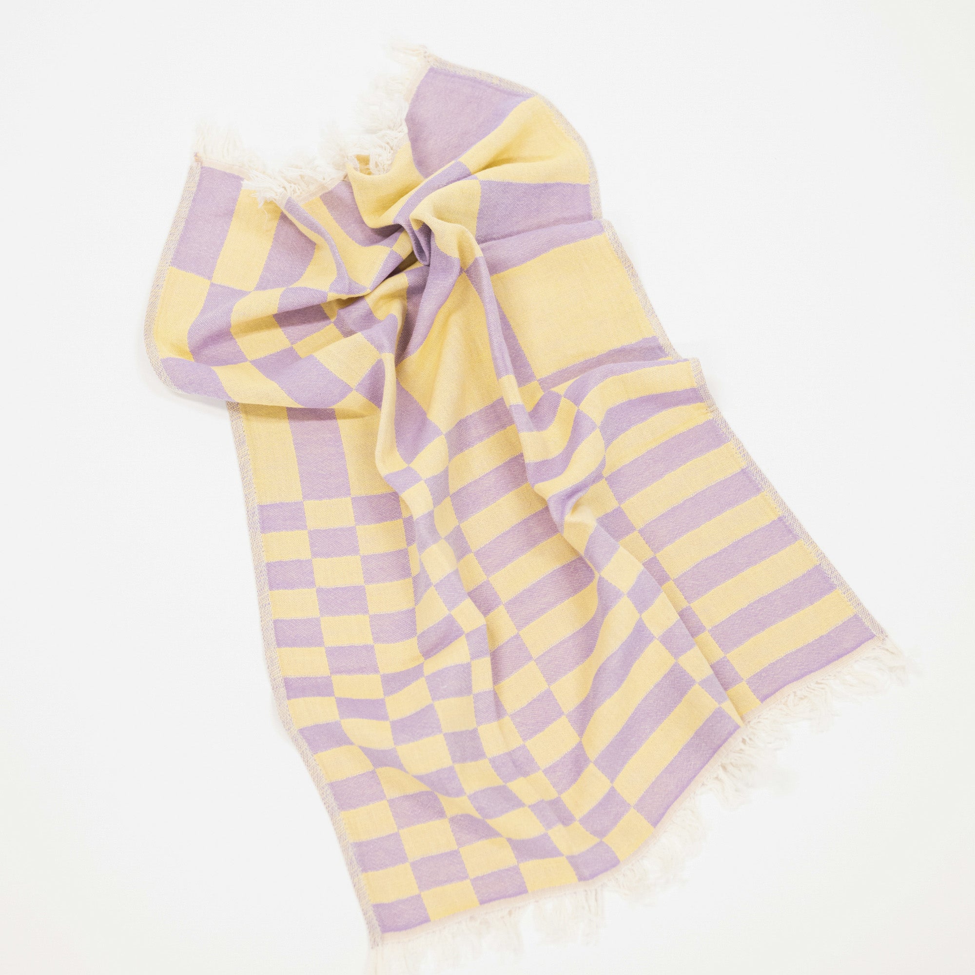A woven kitchen towel with a yellow and lavender checkerboard pattern and fringed edges, laid out on a clean white background.