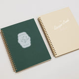 Two spiral-bound recipe notebooks, one green and one beige, on a light gray background.