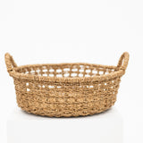 A natural woven seagrass basket with two loop handles, displayed on a white pedestal against a white background.