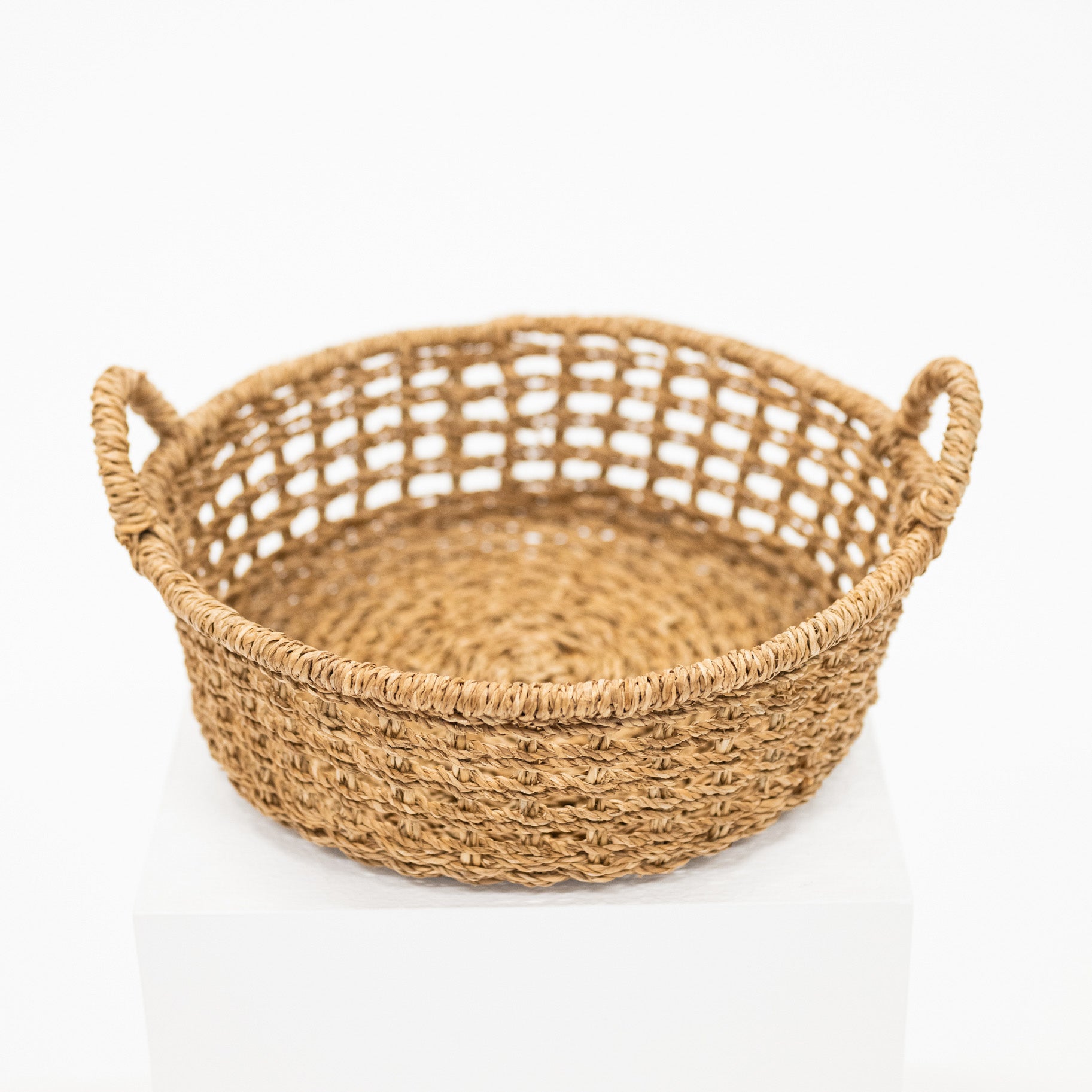 A round woven natural fiber basket with two loop handles, displayed on a white pedestal against a white background.