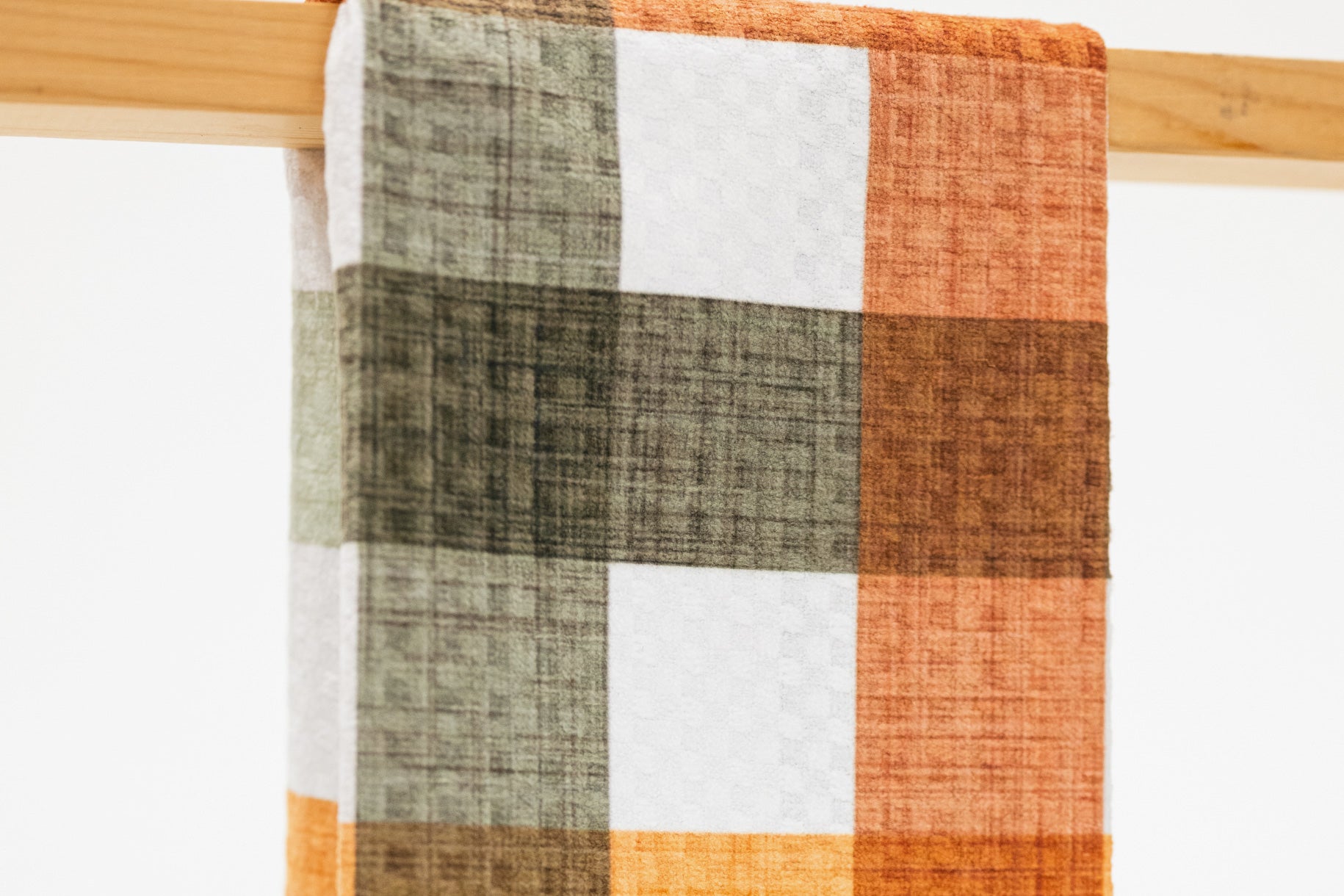 A close-up of a plaid kitchen towel, hanging from a wooden rod. The pattern features blocks of rust orange, dark brown, olive green, and white, with a visible linen-like texture.