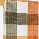 A close-up of a plaid kitchen towel, hanging from a wooden rod. The pattern features blocks of rust orange, dark brown, olive green, and white, with a visible linen-like texture.