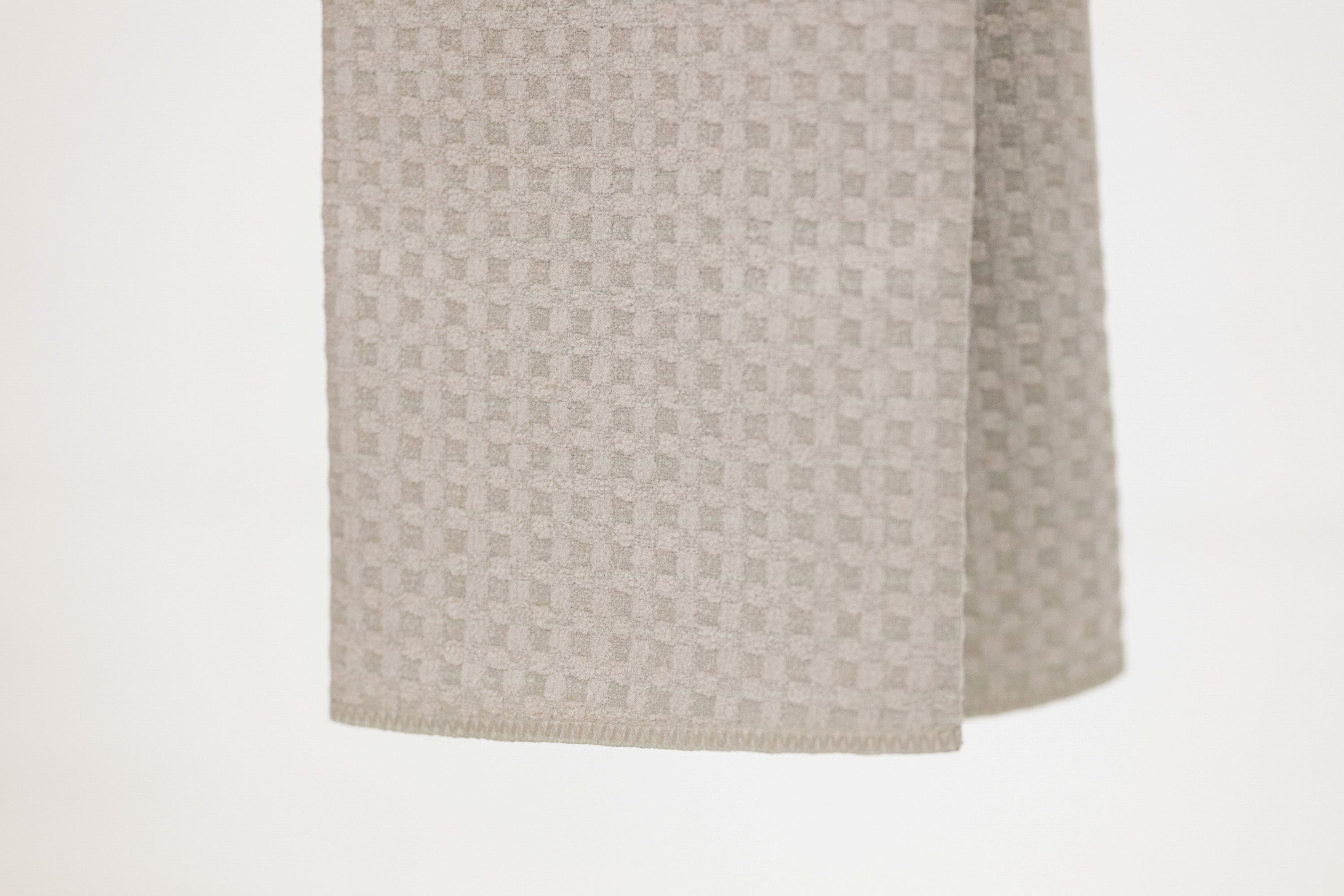 A close-up of a light beige woven fabric with a subtle geometric pattern, hanging against a plain white background.