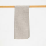 A light beige textured towel with a repeating geometric pattern, hanging from a light wood rod against a solid white background.