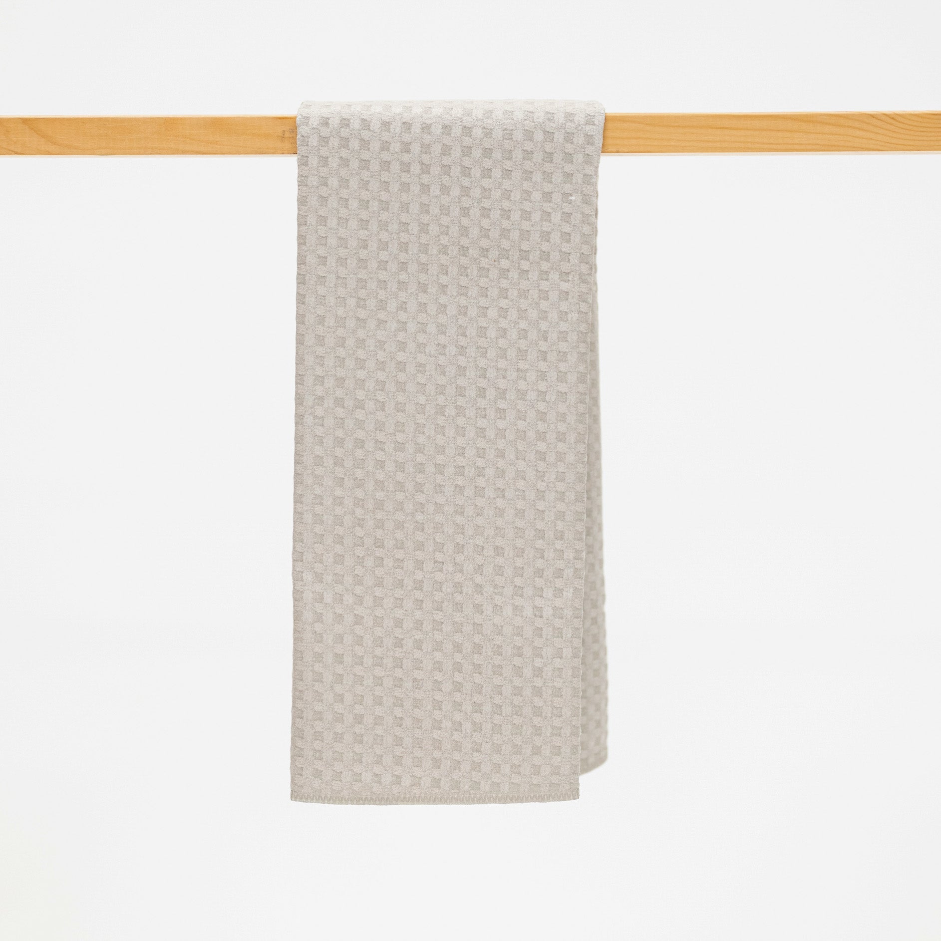 A light beige textured towel with a repeating geometric pattern, hanging from a light wood rod against a solid white background.