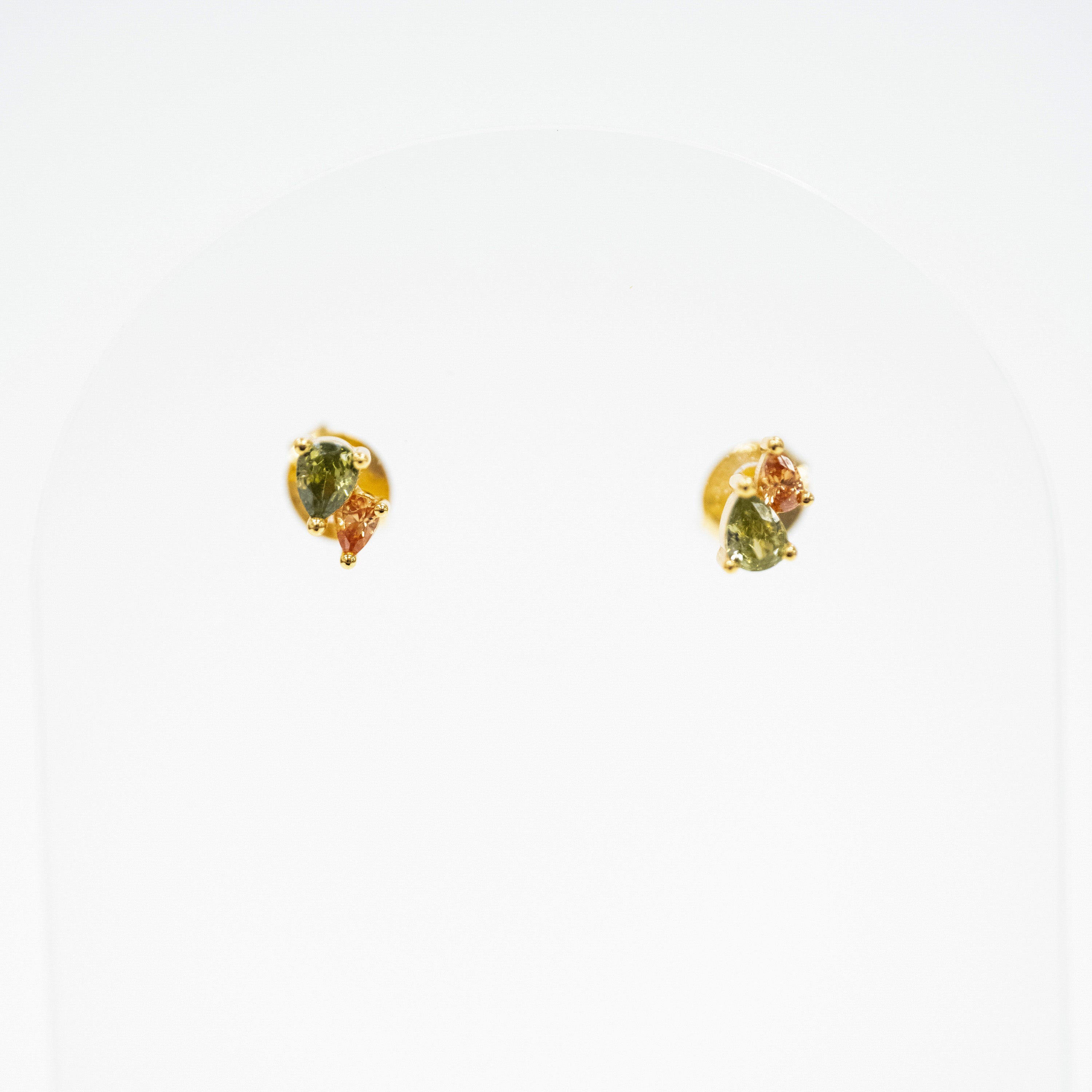 A pair of small gold stud earrings with green and orange stones, displayed on a white arched jewelry stand.