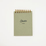 A small square notebook with a muted sage green cover and gold spiral binding, featuring the text 'Elkader' and 'IOWA' printed on the front, set against a plain white background.