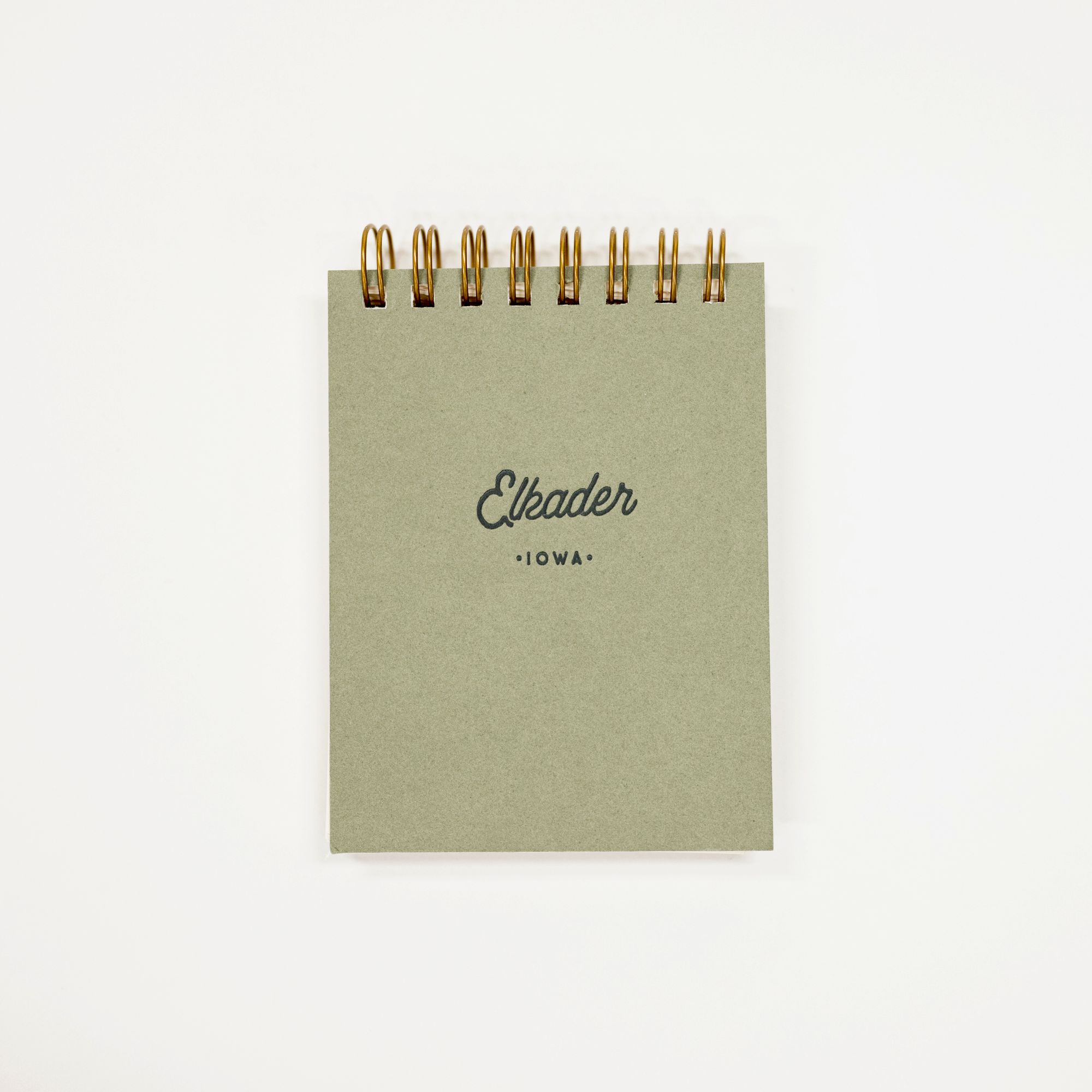 A small square notebook with a muted sage green cover and gold spiral binding, featuring the text 'Elkader' and 'IOWA' printed on the front, set against a plain white background.
