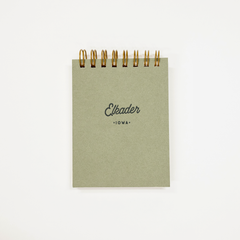 A small square notebook with a muted sage green cover and gold spiral binding, featuring the text 'Elkader' and 'IOWA' printed on the front, set against a plain white background.