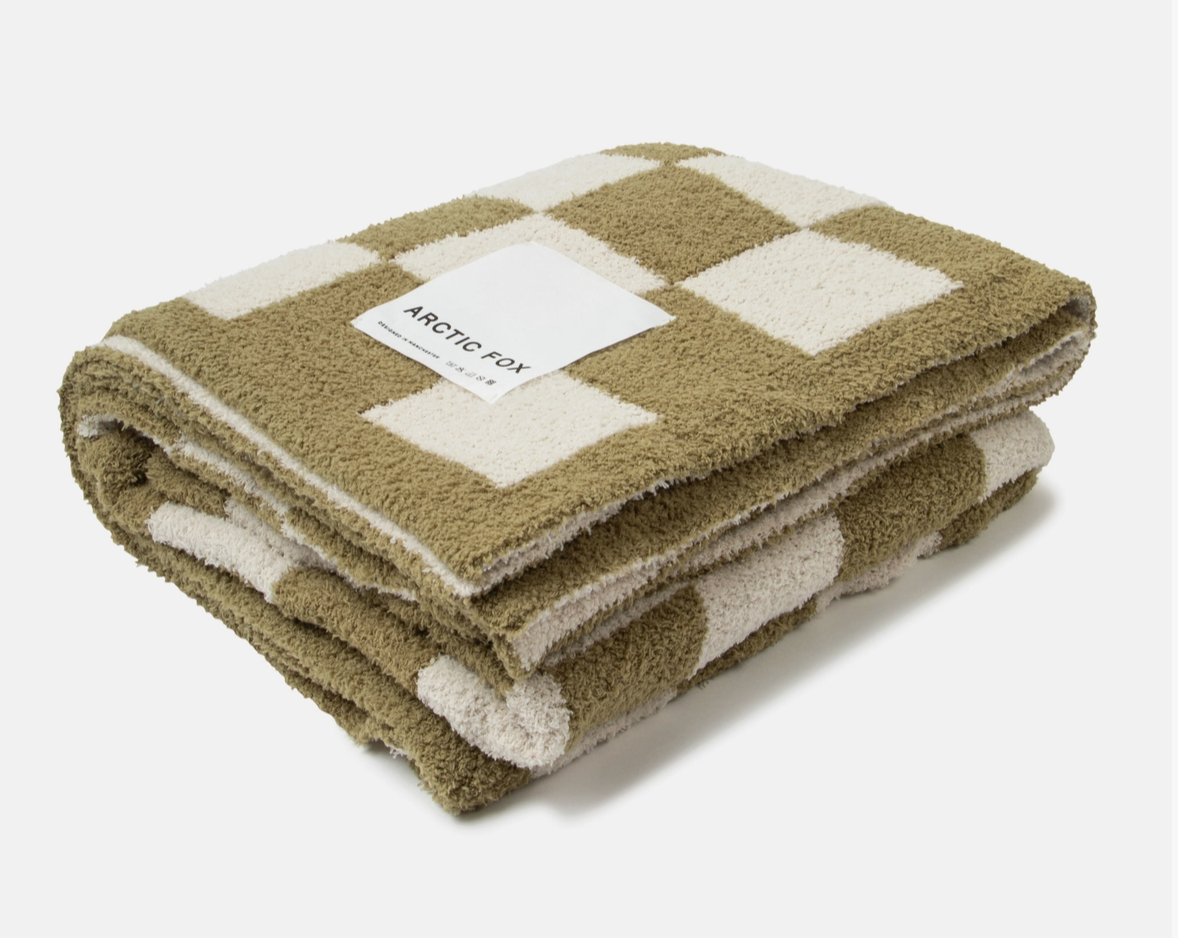 A folded fleece throw blanket with a checkered pattern in olive green and cream, featuring a white paper label that reads 'ARCTIC FOX'.