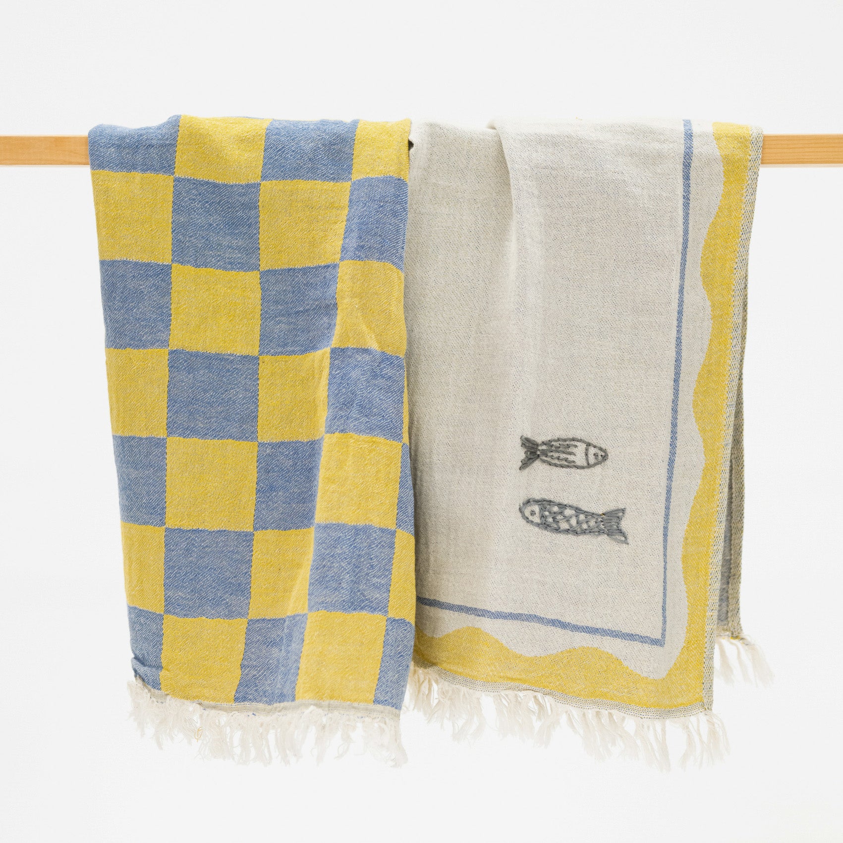 Two woven towels hanging from a wooden rod against a white background. One towel has a yellow and blue checkerboard pattern, and the other is off-white with two fish illustrations and yellow border details.