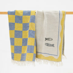 Two woven towels hanging from a wooden rod against a white background. One towel has a yellow and blue checkerboard pattern, and the other is off-white with two fish illustrations and yellow border details.