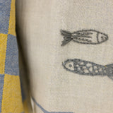 Close-up of a woven textile with two embroidered fish motifs on a light beige background, next to a blue and yellow checkered fabric.