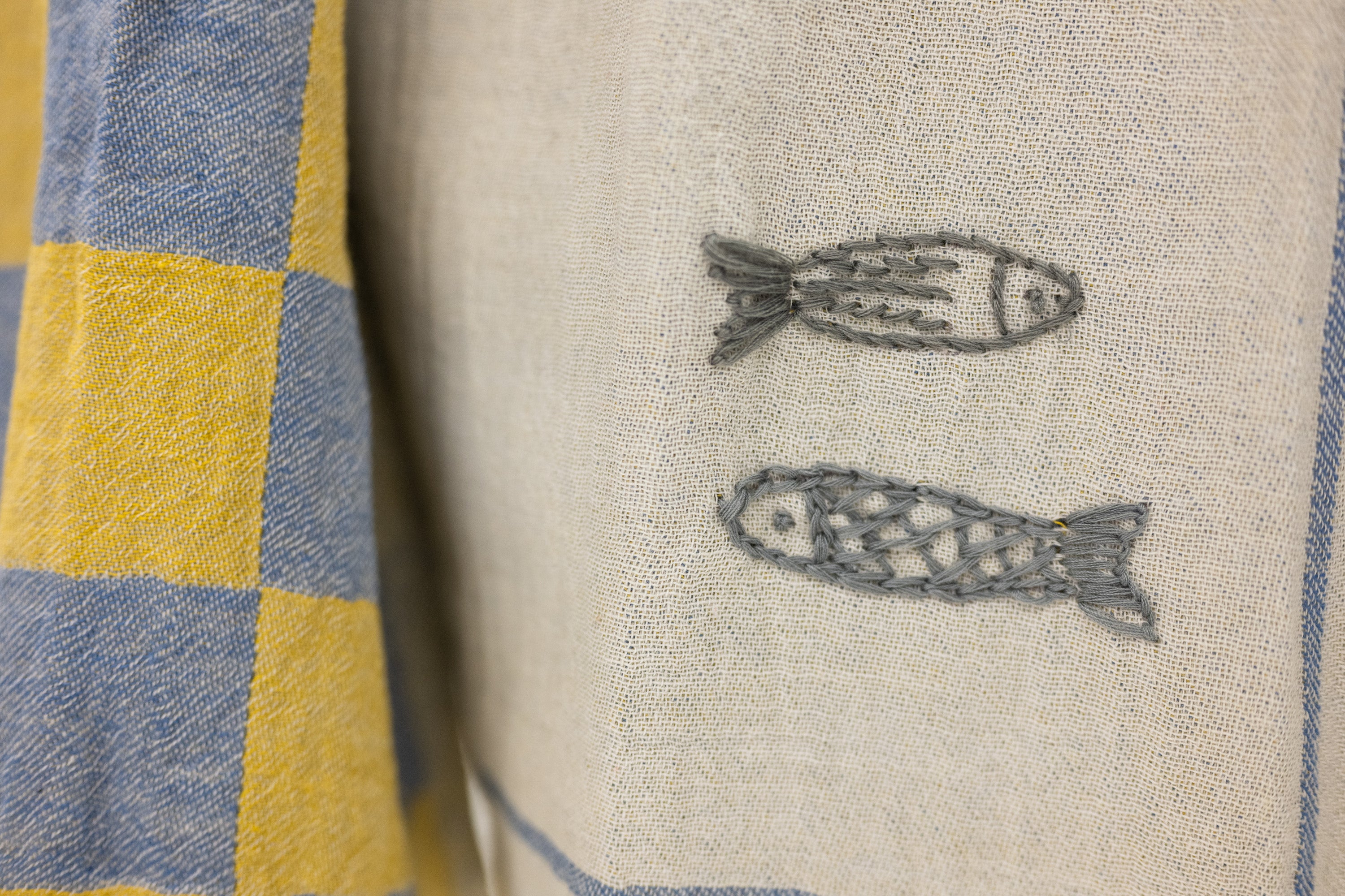 Close-up of a woven textile with two embroidered fish motifs on a light beige background, next to a blue and yellow checkered fabric.