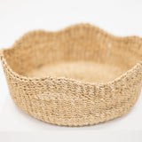 A round woven basket made of natural jute or seagrass with a scalloped rim, displayed on a white pedestal against a clean white background.