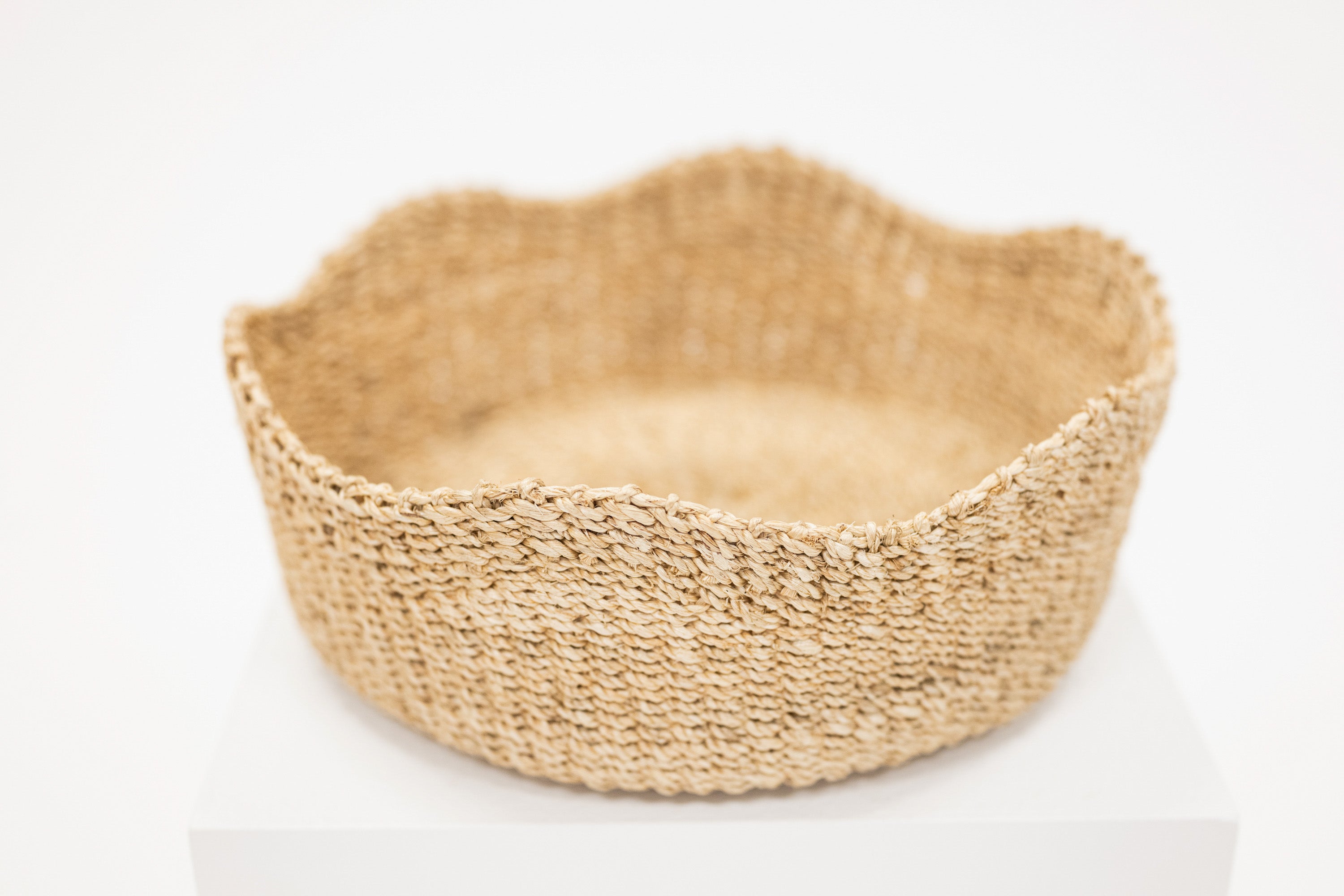 A round woven basket made of natural jute or seagrass with a scalloped rim, displayed on a white pedestal against a clean white background.