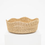 A natural woven seagrass basket with a scalloped rim, displayed on a white pedestal against a clean white background.