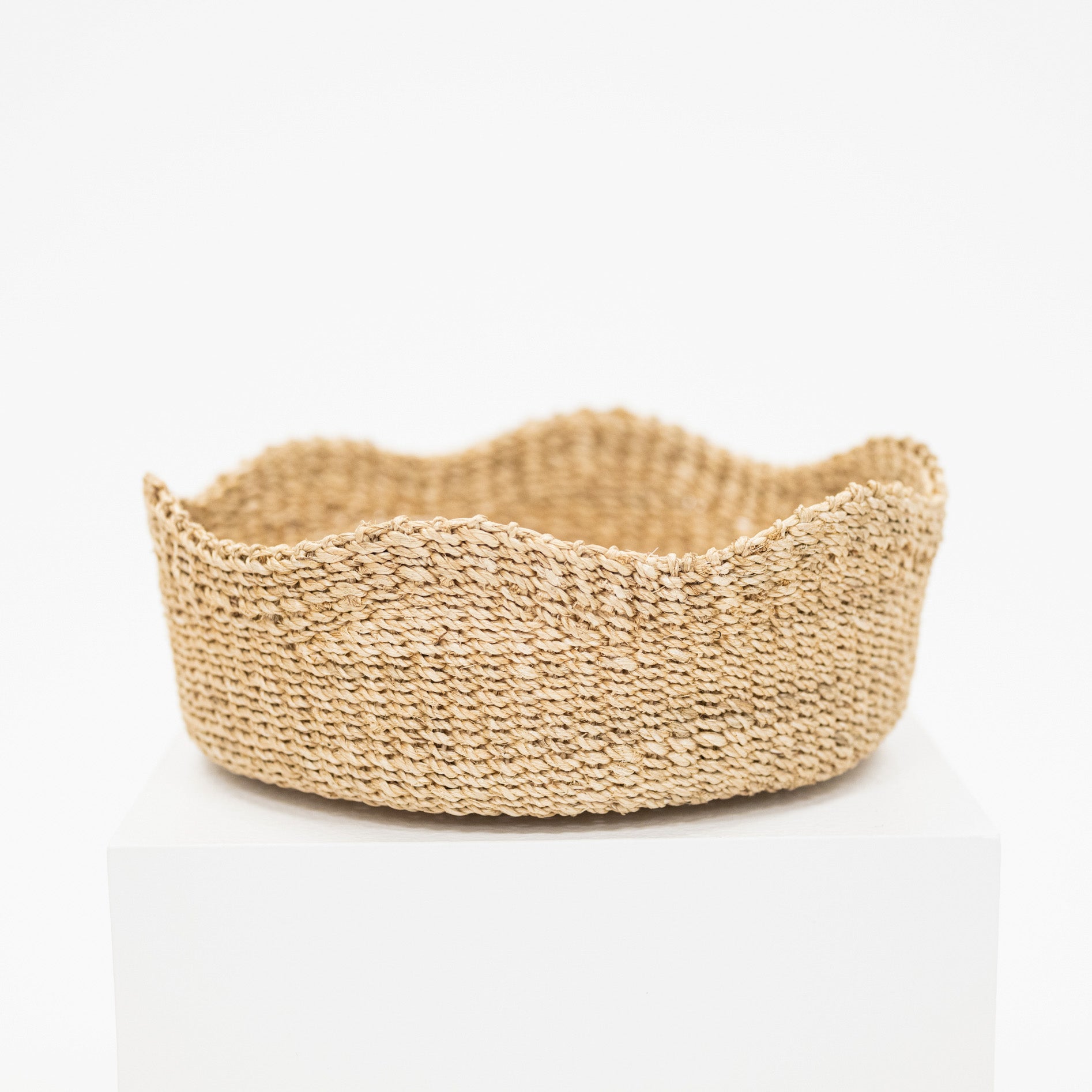 A natural woven seagrass basket with a scalloped rim, displayed on a white pedestal against a clean white background.