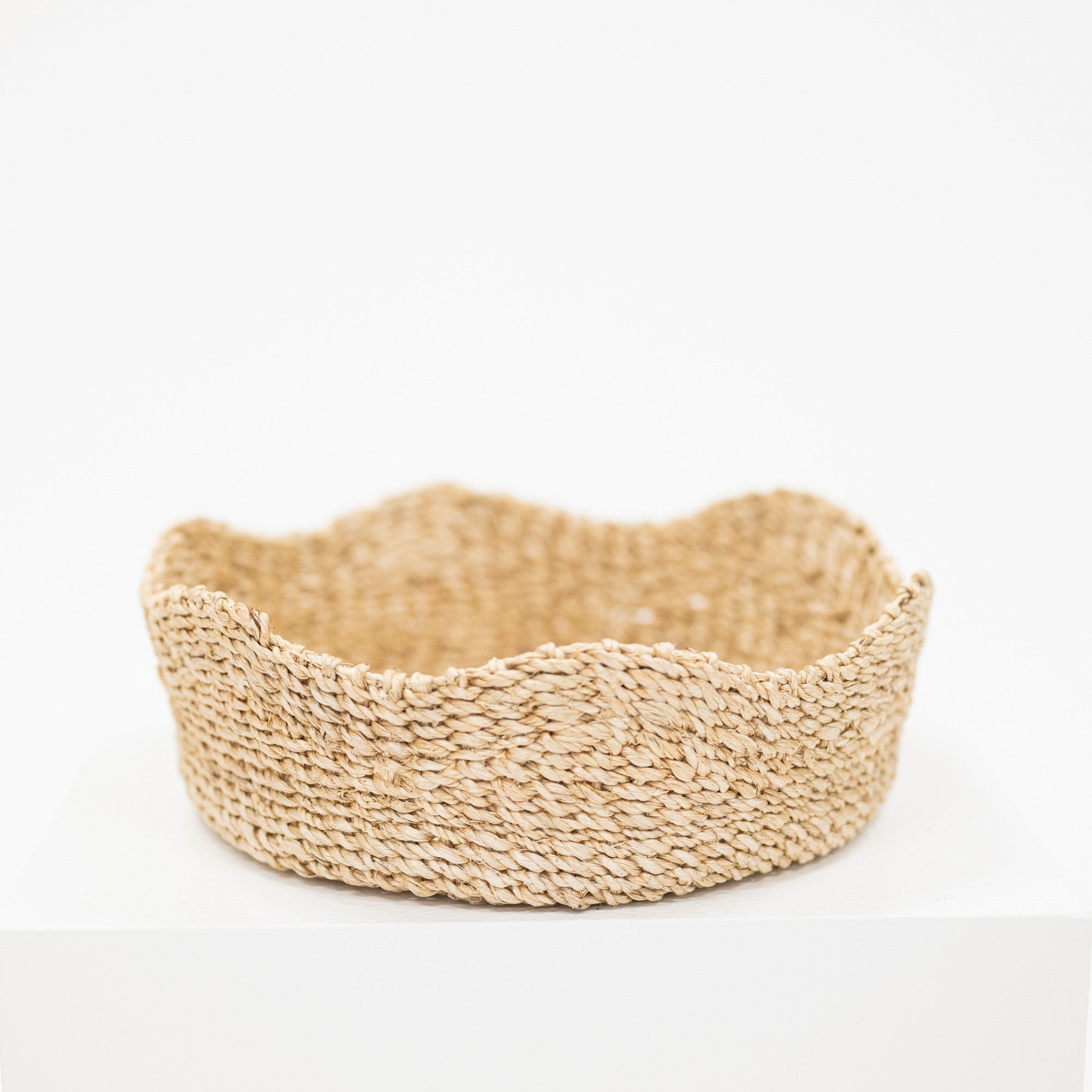 A shallow, scalloped-edge woven basket made of natural straw or seagrass, displayed on a white pedestal against a white background.