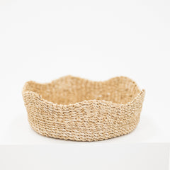 A shallow, scalloped-edge woven basket made of natural straw or seagrass, displayed on a white pedestal against a white background.