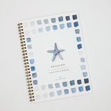 A spiral-bound watercolor workbook titled 'SEASIDE watercolor workbook' on a white cover, decorated with a blue starfish in the center and a border of blue and gray watercolor squares, photographed on a light gray background.