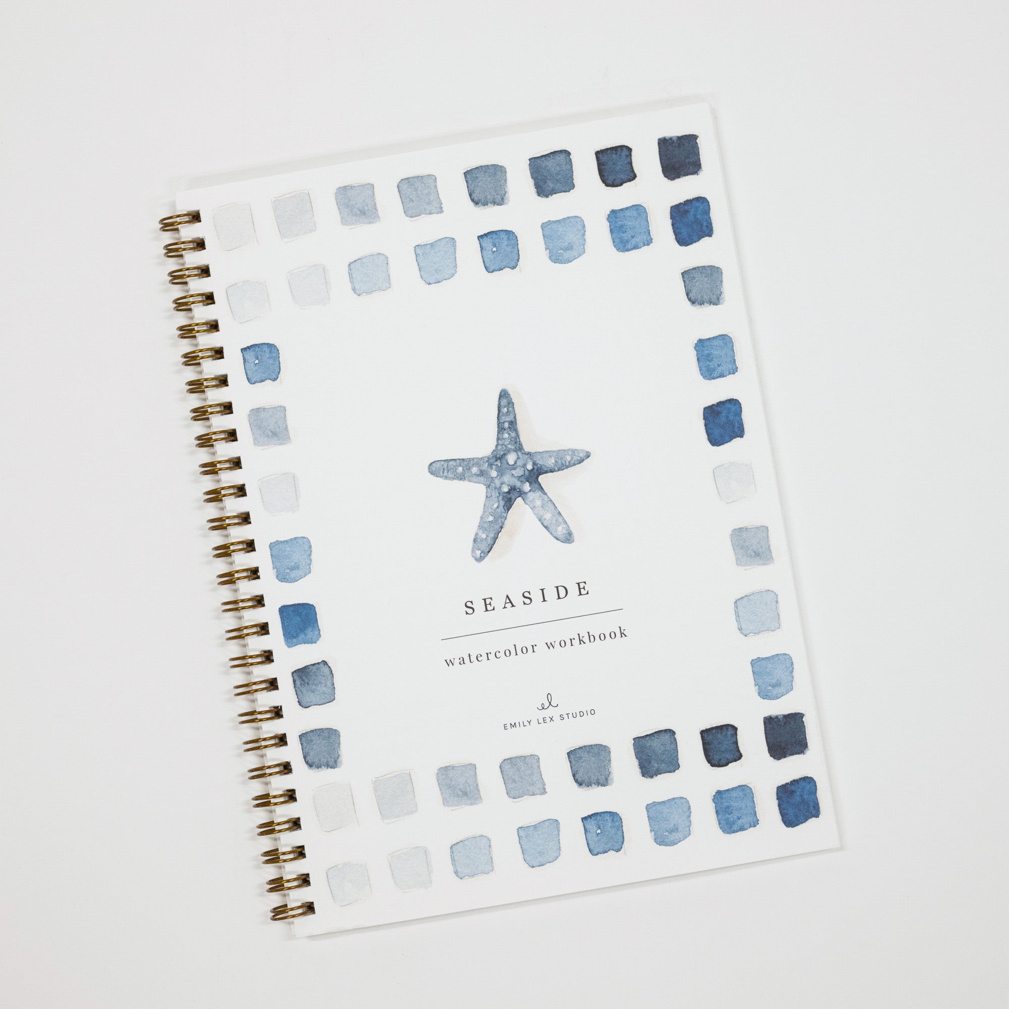A spiral-bound watercolor workbook titled 'SEASIDE watercolor workbook' on a white cover, decorated with a blue starfish in the center and a border of blue and gray watercolor squares, photographed on a light gray background.
