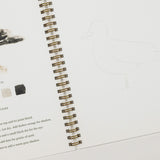 An open spiral-bound art journal or sketchbook showing a bird illustration, a color palette section with paint swatches, and instructions, alongside a faint line drawing of a bird on the right page.