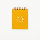 A small spiral-bound notebook with a bright yellow cover featuring a simple white line drawing of a smiling sun, photographed on a clean white background.