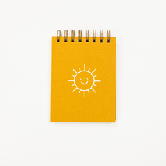 A small spiral-bound notebook with a bright yellow cover featuring a simple white line drawing of a smiling sun, photographed on a clean white background.