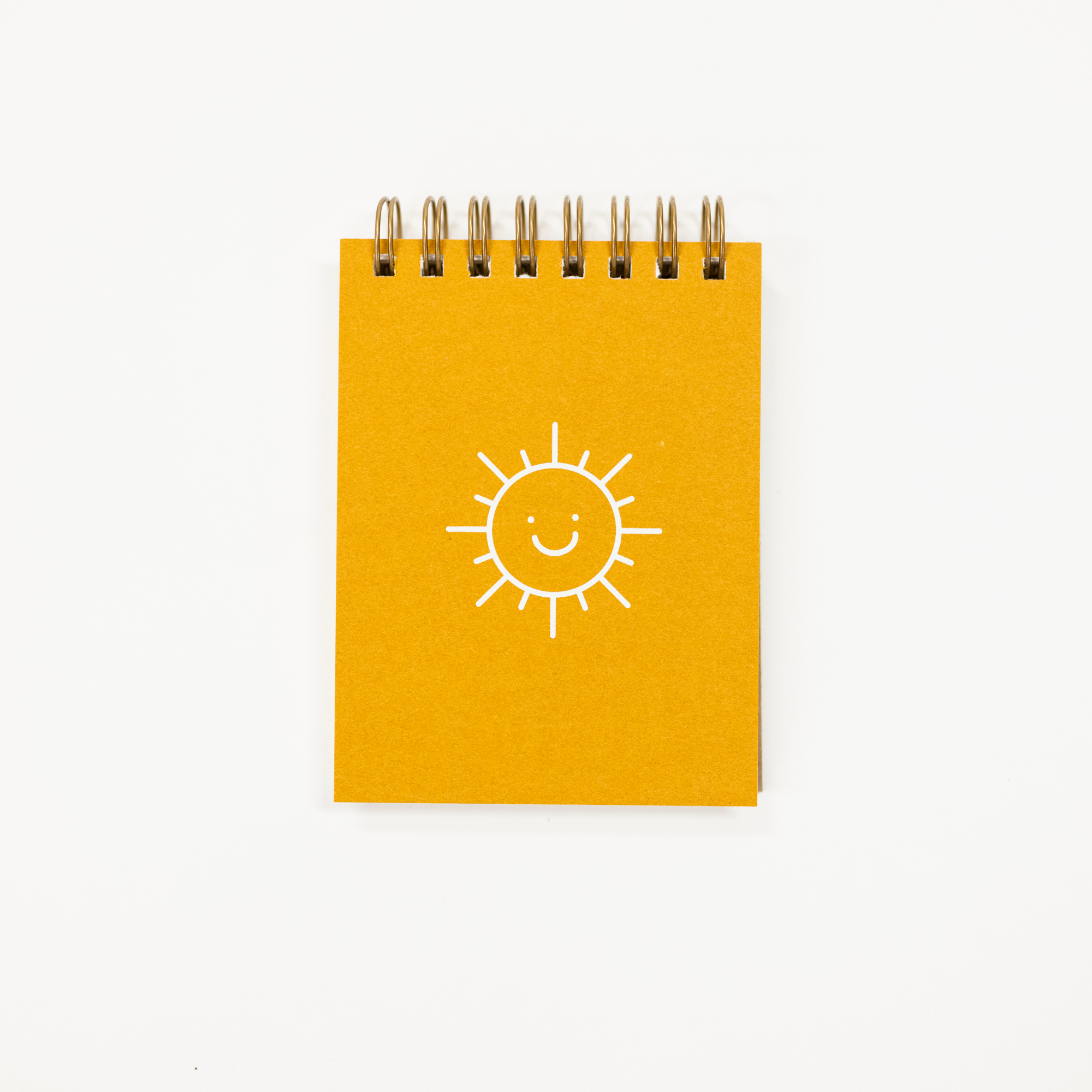 A small spiral-bound notebook with a bright yellow cover featuring a simple white line drawing of a smiling sun, photographed on a clean white background.