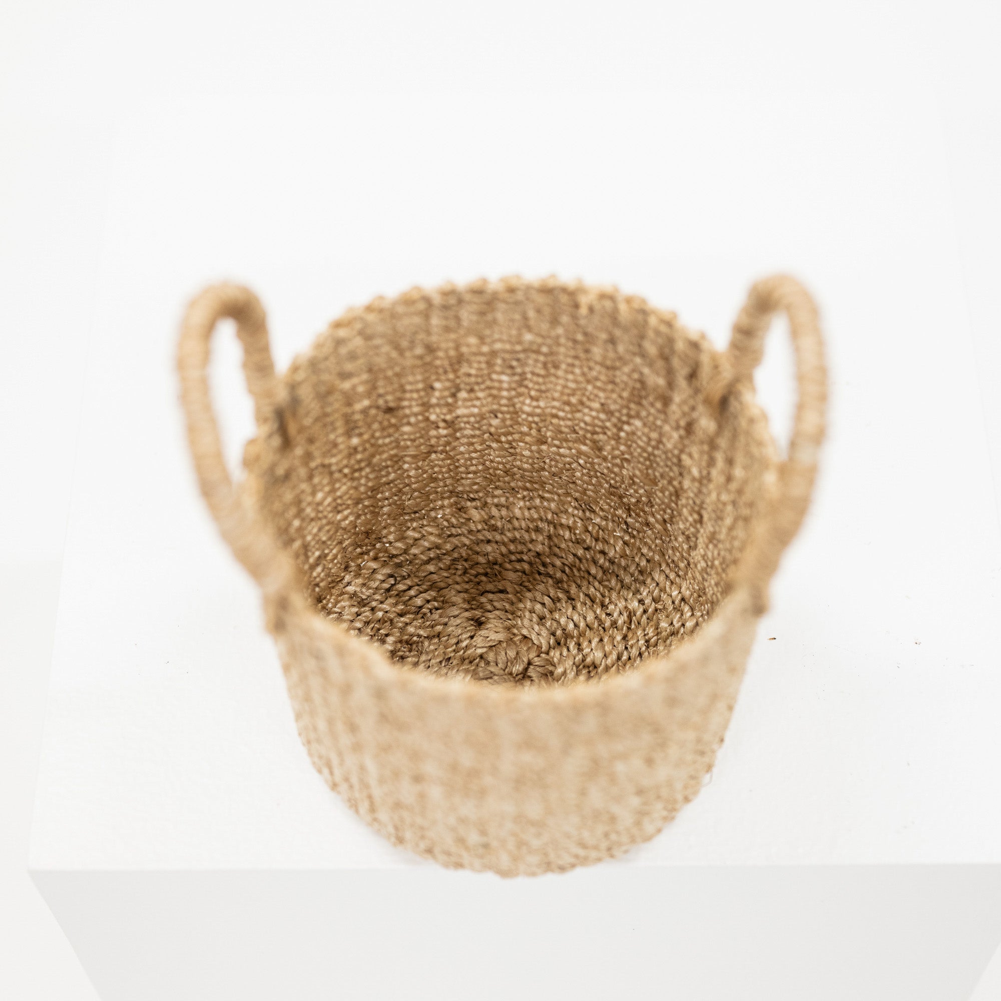 A natural woven basket with two loop handles, made from tan jute or seagrass, photographed from a top-down angle on a white platform against a white background.