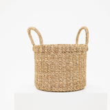 A natural woven seagrass basket with two loop handles, displayed on a white pedestal against a clean white background.