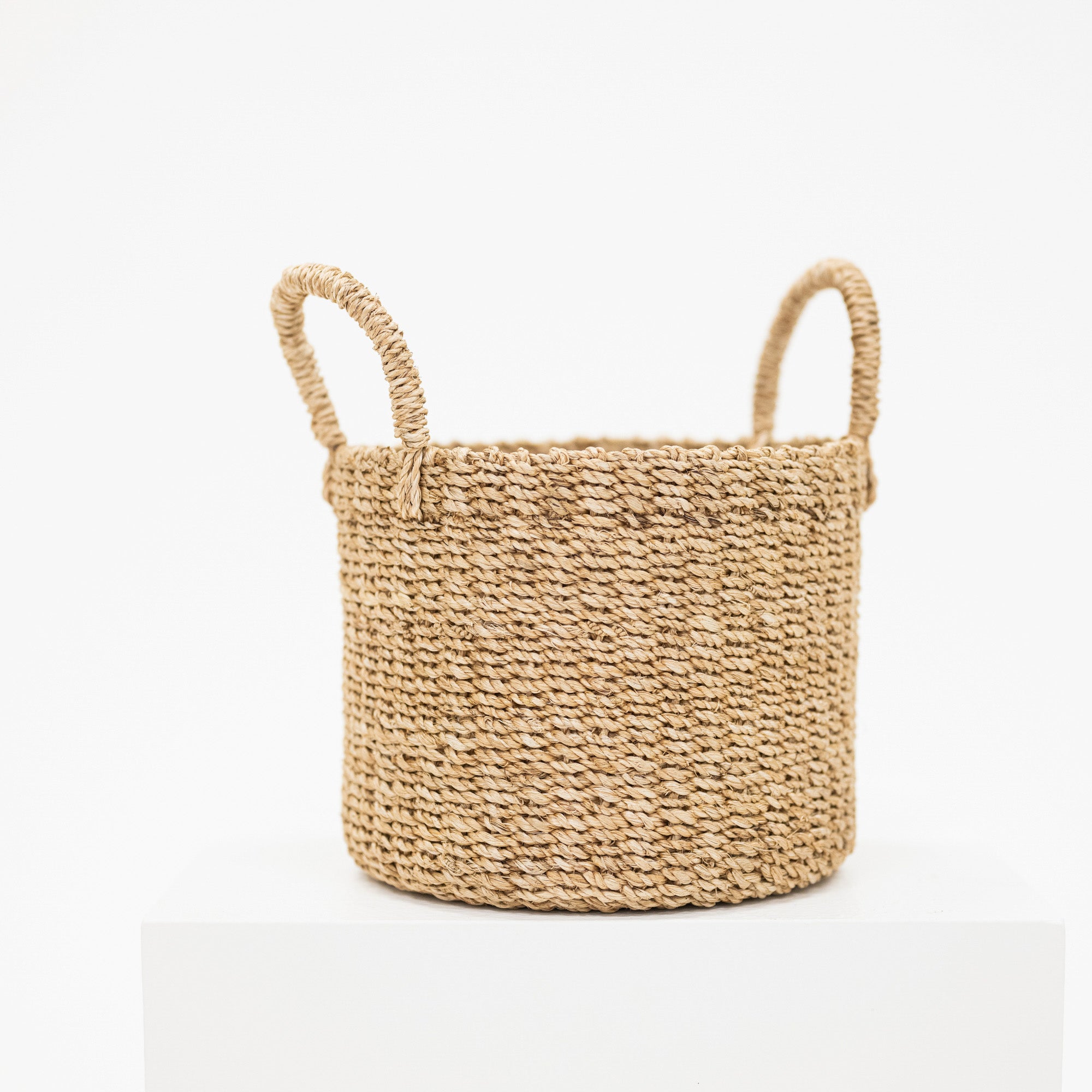 A natural woven seagrass basket with two loop handles, displayed on a white pedestal against a clean white background.
