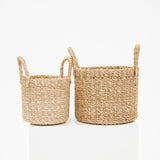 Two natural woven seagrass storage baskets with loop handles, shown in two different sizes on a white reflective surface against a white background.