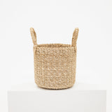 A small woven basket made of natural beige seagrass or straw, featuring two loop handles, displayed on a white pedestal against a clean white background.