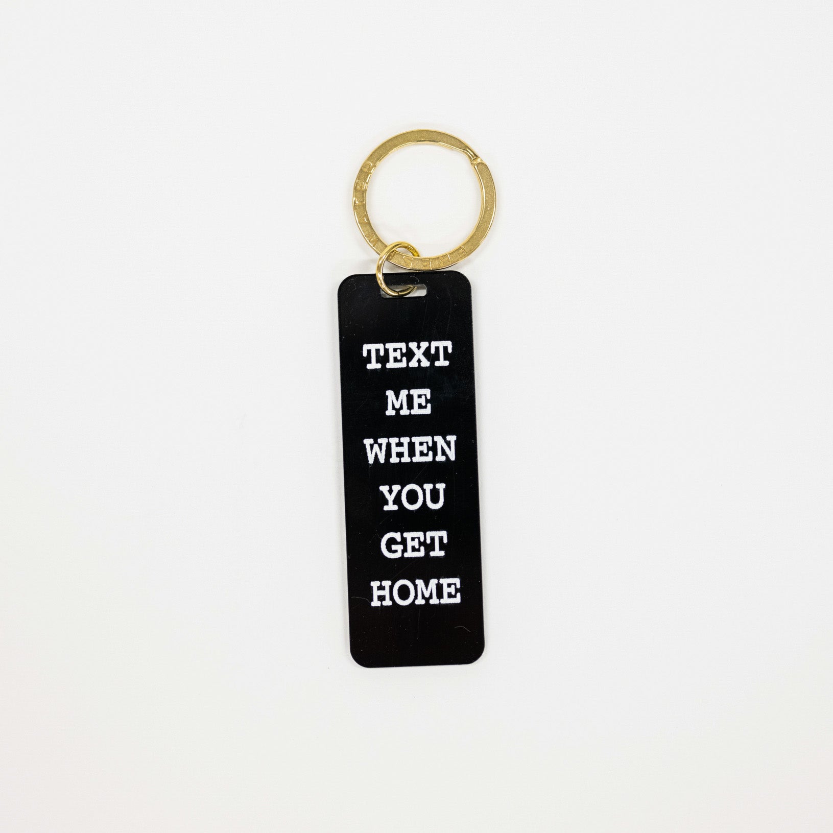A black rectangular acrylic keychain with white text reading 'TEXT ME WHEN YOU GET HOME' attached to a gold split keyring, shown on a plain white background.